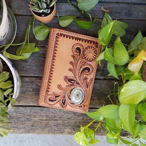 May include: A tan leather wallet with intricate floral tooling and a silver concho. The wallet is lying on a wooden surface with green plants.