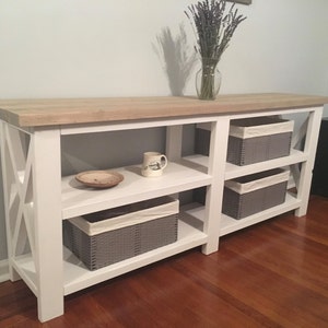 May include: White console table with a wood top and three shelves. The table has an X-shaped design on the front and sides. There are two gray baskets on the bottom shelf, one gray basket on the middle shelf, and a white mug on the top shelf.