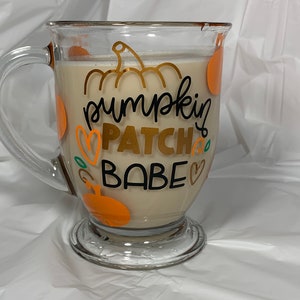 May include: A clear glass mug with a handle, filled with a light brown beverage. The mug has a pumpkin-themed design with the text "pumpkin patch babe" in black and gold lettering.