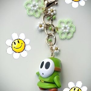 Könnte beinhalten: Ein grüner Shy Guy-Schlüsselanhänger mit weißer Maske, braunen Schuhen und einem goldfarbenen Verschluss. Der Schlüsselanhänger ist mit kleinen weißen und grünen Blumenanhängern und Perlenakzenten verziert. Smiley-Blumenaufkleber befinden sich im Hintergrund.
