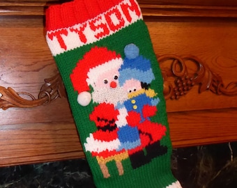 May include: A green, red, and white knitted Christmas stocking with a Santa Claus and a child figure embroidered on the front. The stocking has the word "TYSON" knitted into the top.