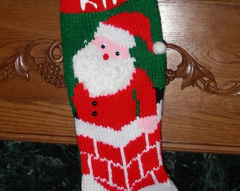 May include: A green, white, and red knitted Christmas stocking with a Santa Claus design. The stocking has the name "KIA" written on it.
