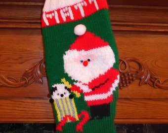 May include: A green, white, and red knitted Christmas stocking with a Santa Claus and a toy figure. The stocking has a white pom-pom on top.