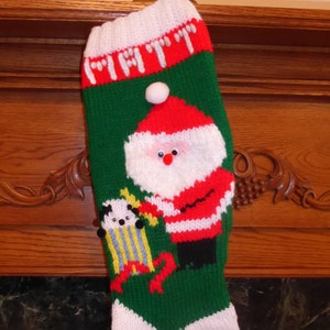 May include: A green, white, and red knitted Christmas stocking with a Santa Claus and a toy figure. The stocking has a white pom-pom on top.