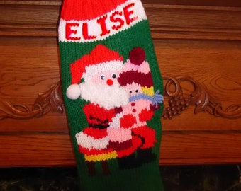 May include: A green, red, and white knitted Christmas stocking with the name "Elise" embroidered on the front. The stocking features a Santa Claus and a snowman design.