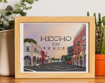 May include: A framed print with the text "HECHO EN VENICE" depicts a street scene with buildings and a road. The print is in a light wood frame and is displayed on a wooden surface with plants and a camera.