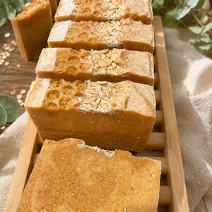 May include: Close-up of several rectangular bars of handmade soap. The soaps are light brown with textured surfaces, some with honeycomb and oat embellishments. They are arranged on a wooden tray, suggesting a natural, organic theme.