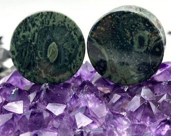 May include: Two round, green stone plugs with a marbled pattern. The plugs are displayed on a bed of purple amethyst crystals. The plugs have a flat, circular face and a cylindrical body.