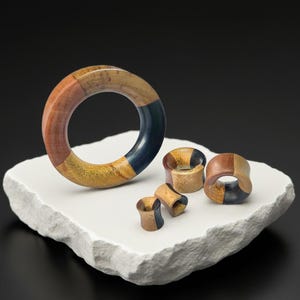 May include: A collection of wooden ear plugs and a large ring, displayed on a white, textured surface against a black background. The plugs and ring are made of various wood tones, including brown, yellow, and blue, creating a geometric pattern.