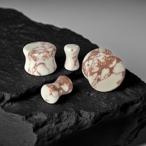 May include: Four white and brown marbled stone ear plugs of varying sizes are displayed on a dark, textured surface. The plugs have a unique pattern and shape, showcasing a natural, organic aesthetic. The plugs are designed for body modification.