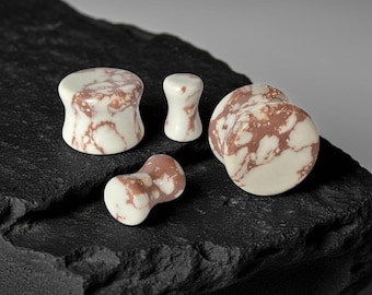 May include: A set of four ear plugs featuring a white and dusty rose marbled pattern. The plugs have a smooth, cylindrical shape with flared ends, showcasing a distinct veined texture across the surface of each individual piece.