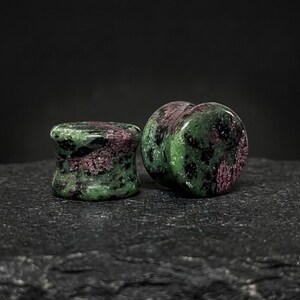 May include: A pair of circular ear plugs crafted from zoisite stone, featuring a polished surface with a mottled pattern of deep forest green, vibrant magenta, and black inclusions. The plugs have a concave, flared shape for secure placement.