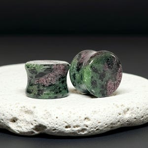 May include: A pair of cylindrical ear plugs crafted from zoisite stone, featuring a mottled pattern of deep forest green, vibrant magenta, and black inclusions. The plugs have a smooth, polished finish with a concave center and flared edges.