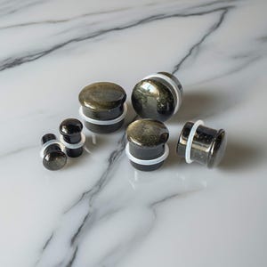May include: A collection of black and gold plugs with white O-rings. The plugs vary in size and shape, with a polished, reflective surface. The jewelry is displayed on a white and gray marble surface.