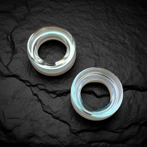 May include: Two clear, circular ear gauges with iridescent highlights sit on a dark, textured surface. The gauges have a layered design, with a central hole and a rounded outer edge. The light catches the edges, creating a subtle glow.
