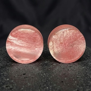 May include: A pair of circular ear plugs featuring a translucent, pale pink hue with internal swirling patterns and a smooth, polished finish. The plugs have a flat front surface and a consistent, rounded shape, showcasing natural-looking inclusions.