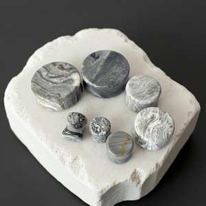 May include: A collection of gray and white stone ear plugs of various sizes. The plugs have a marbled appearance, with swirling patterns in shades of gray and white. They are displayed on a white, textured surface.