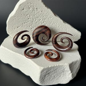 May include: A collection of six wooden spiral-shaped ear gauges in varying sizes. The gauges are a rich, dark brown color with visible wood grain. They are displayed on a textured white surface against a dark background.