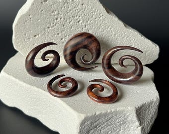 May include: A collection of six wooden spiral-shaped ear gauges in varying sizes. The gauges are a rich, dark brown color with visible wood grain. They are displayed on a textured white surface against a dark background.