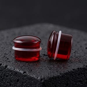 May include: A pair of deep red, translucent glass ear plugs with a smooth, rounded front surface and a clear silicone O-ring positioned around the center of each cylindrical body, designed for stretched ear piercings with a polished finish.