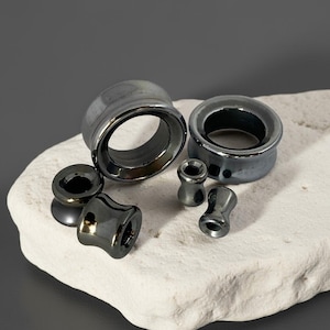 May include: An assortment of dark gray ear gauges in various sizes and shapes. The collection includes flared plugs, saddle plugs, and small, cylindrical gauges. The gauges are displayed on a white, textured surface against a gray backdrop.