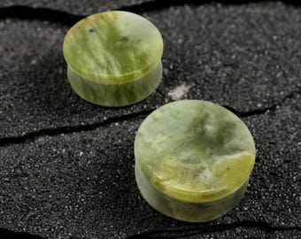 May include: Two green jade stone ear plugs. The plugs are round with a flat face and a slightly rounded back. The jade has a marbled appearance with shades of green and yellow. The plugs are displayed on a dark, textured surface.