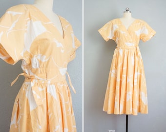 May include: A yellow wrap dress with a white floral print. The dress has short sleeves and a V-neckline. It is tied at the waist with a sash.