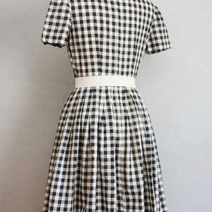 May include: A black and white checkered dress with short sleeves and a white belt. The dress is displayed on a mannequin and features a pleated skirt. The fabric has a classic gingham pattern.