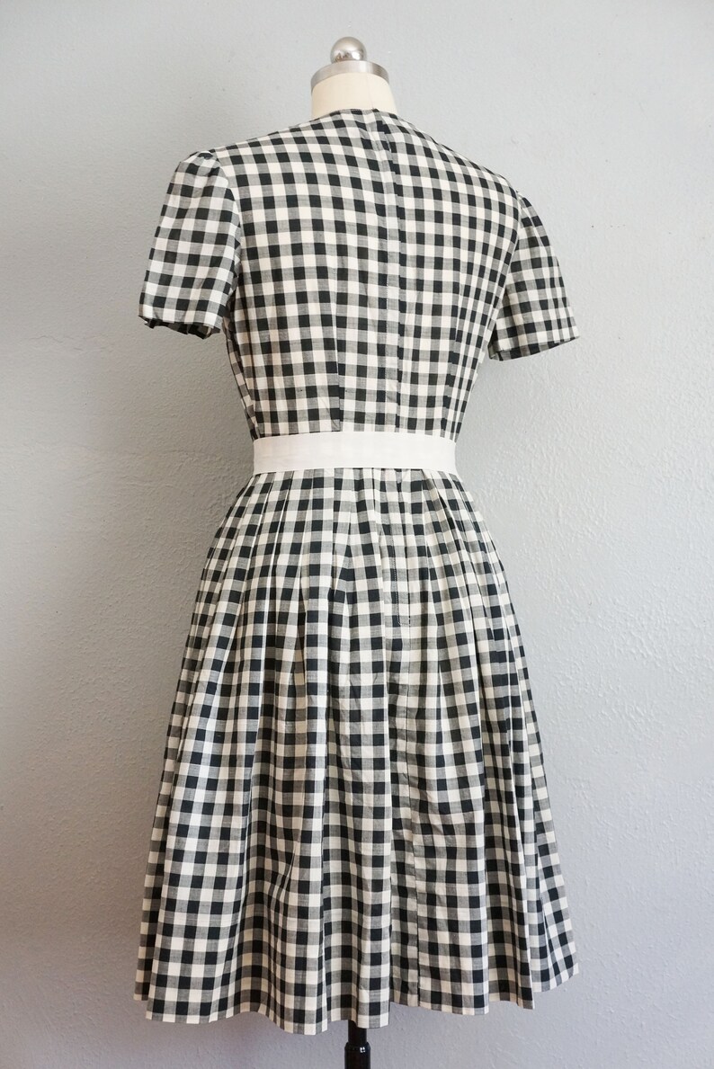 May include: A black and white checkered dress with short sleeves and a white belt. The dress is displayed on a mannequin and features a pleated skirt. The fabric has a classic gingham pattern.