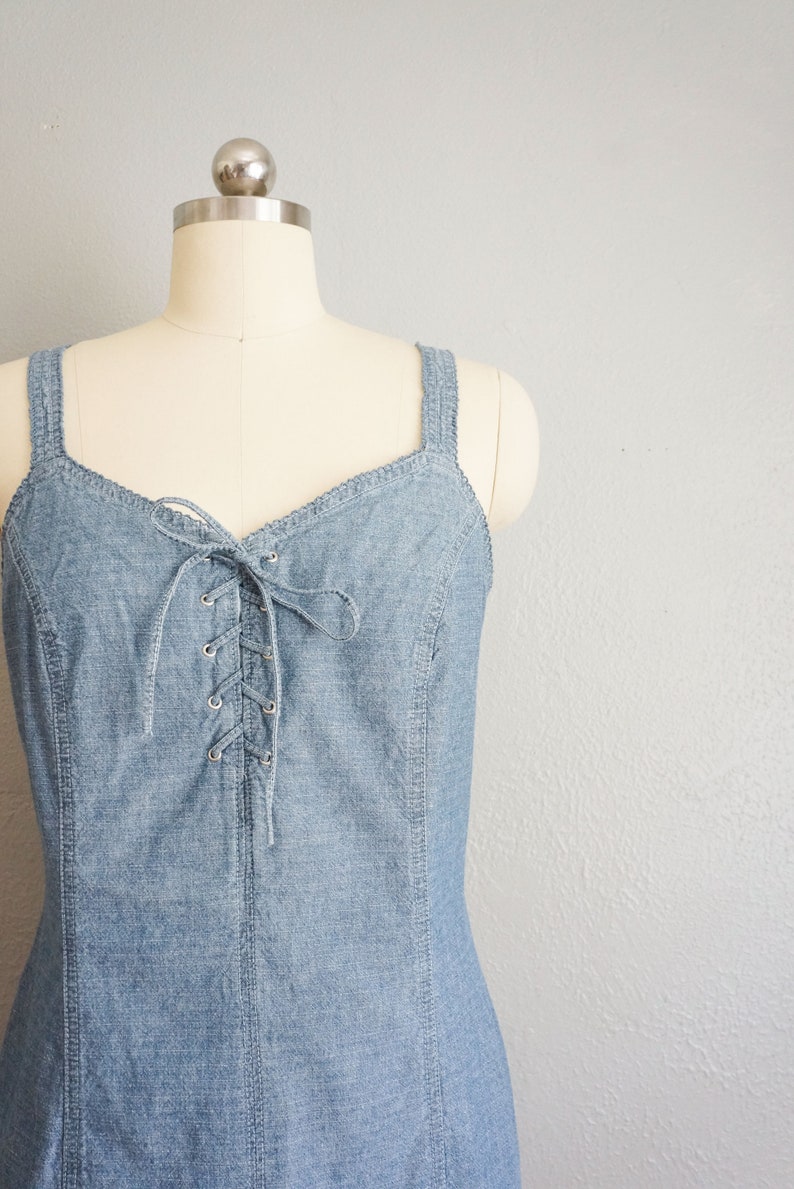May include: A light blue denim dress with a lace-up front and thin straps. The dress is on a white mannequin.