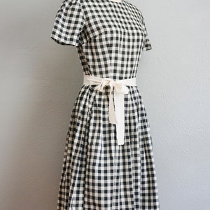 May include: A black and white gingham dress with short sleeves and a cream-colored tie belt. The dress is displayed on a mannequin against a gray background. The dress has a classic, vintage style.