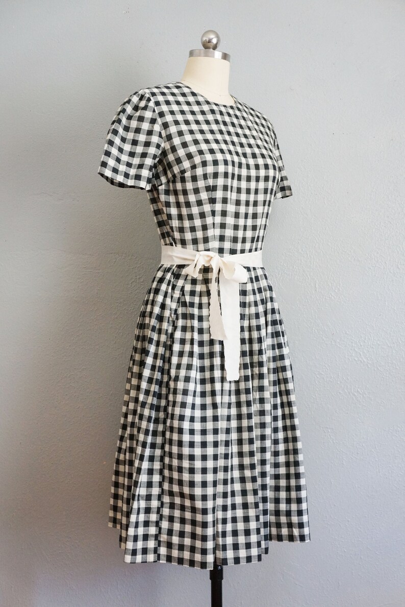 May include: A black and white gingham dress with short sleeves and a cream-colored tie belt. The dress is displayed on a mannequin against a gray background. The dress has a classic, vintage style.