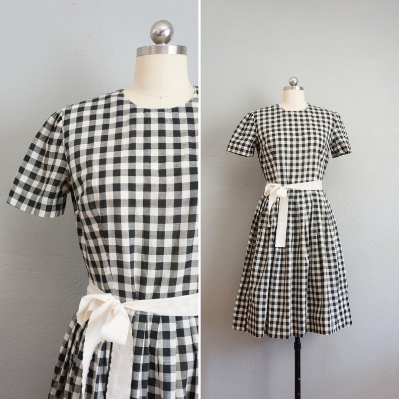 May include: A black and white gingham dress with short sleeves and a white sash tied at the waist. The dress is displayed on a mannequin against a gray background. The dress has a classic, vintage style.