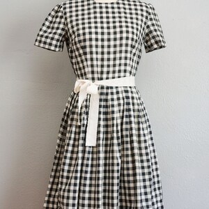 May include: A black and white gingham dress with a white tie belt. The dress has short sleeves and a knee-length skirt. The dress is displayed on a mannequin against a gray background.