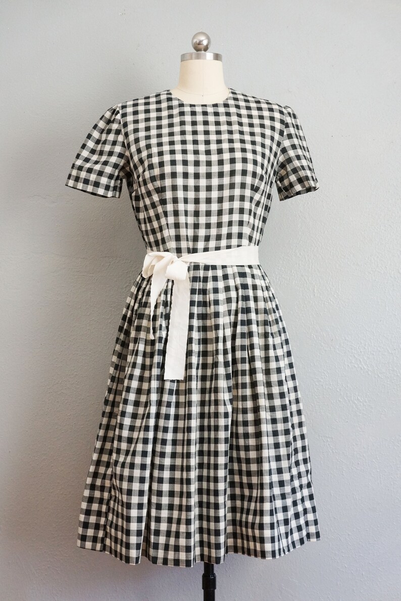 May include: A black and white gingham dress with a white tie belt. The dress has short sleeves and a knee-length skirt. The dress is displayed on a mannequin against a gray background.