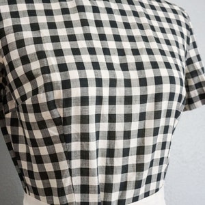 May include: A black and white checkered dress with a white sash tied in a bow. The dress has short sleeves and a fitted bodice. The fabric has a classic gingham pattern.
