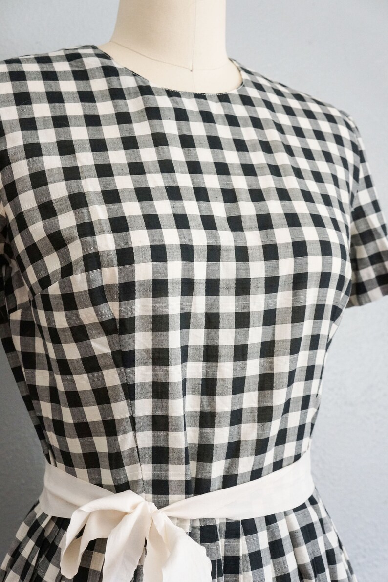 May include: A black and white checkered dress with a white sash tied in a bow. The dress has short sleeves and a fitted bodice. The fabric has a classic gingham pattern.