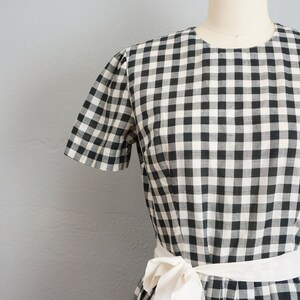 May include: A black and white gingham dress with short sleeves and a white sash tied at the waist. The dress is displayed on a mannequin against a gray background. The fabric has a classic checkered pattern.
