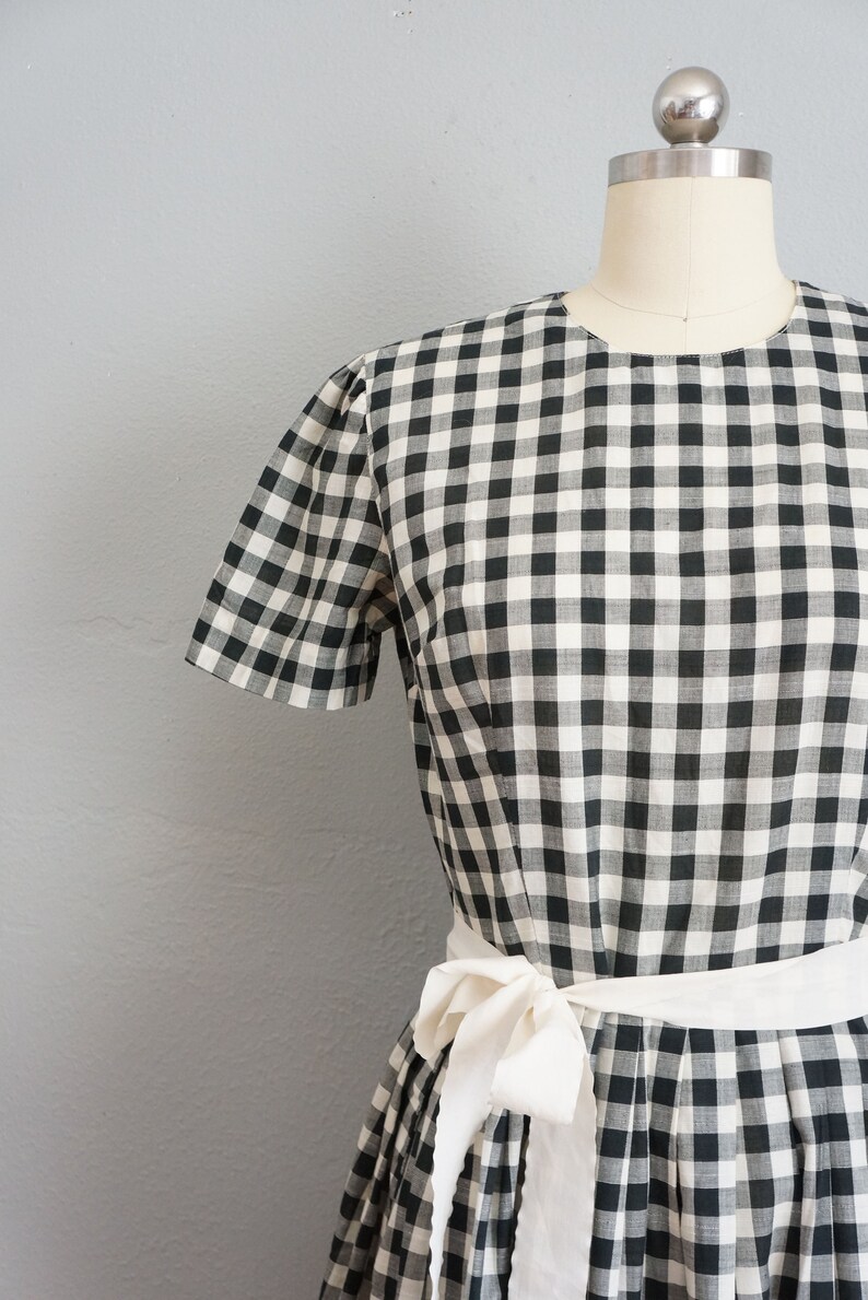 May include: A black and white gingham dress with short sleeves and a white sash tied at the waist. The dress is displayed on a mannequin against a gray background. The fabric has a classic checkered pattern.