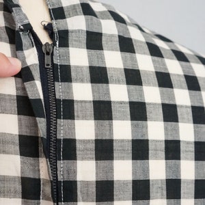 May include: Close-up of a black and white checkered fabric with a zipper. The zipper is dark gray and runs vertically along the fabric. The fabric is a classic gingham pattern, with the checks being a medium size. The garment is on a mannequin.