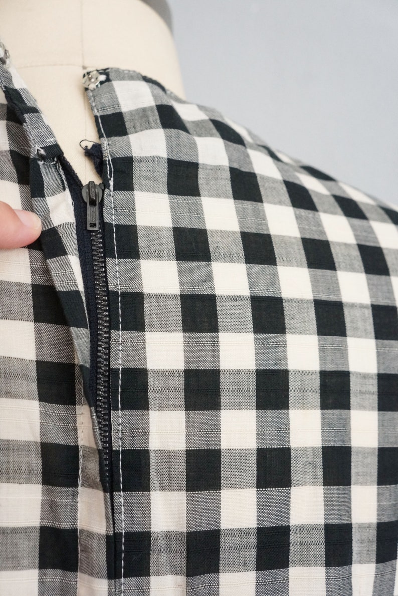 May include: Close-up of a black and white checkered fabric with a zipper. The zipper is dark gray and runs vertically along the fabric. The fabric is a classic gingham pattern, with the checks being a medium size. The garment is on a mannequin.