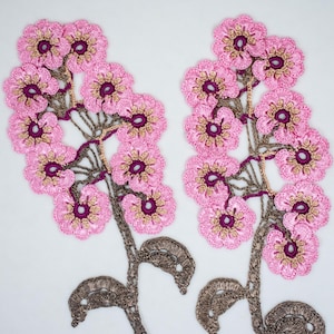 Puede incluir: Dos motivos florales de ganchillo en rosa y marrón. Cada motivo presenta un grupo de flores rosas con centros marrones, conectados por un tallo marrón con hojas.