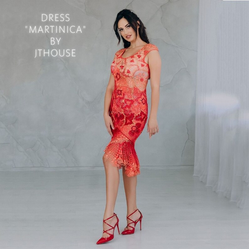 May include: A woman wearing a red and orange crochet dress with a floral pattern. The dress has a fitted bodice and a flared skirt. The dress is labeled "Dress "Martini&ccedil;a" by JTHouse".