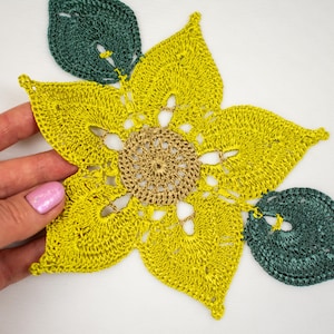 Può includere: Un fiore giallo all'uncinetto con un centro marrone e foglie verdi. Il fiore è realizzato con un delicato motivo a punti e ha un aspetto traforato.