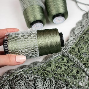 Puede incluir: Primer plano de carretes de hilo verde, parcialmente envueltos en una red, junto a una pieza de ganchillo. El hilo es de un verde oliva oscuro, y el artículo de ganchillo presenta tonos verdes y grises. El fondo es blanco.