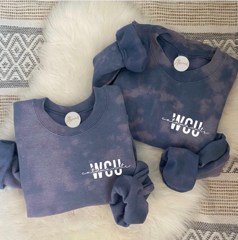 May include: Two blue tie-dye crewneck sweatshirts with the word "WCU" in white script. Each sweatshirt has a small circular tag that says "Athread". One sweatshirt has a matching scrunchie. The sweatshirts are on a white fur-like surface.