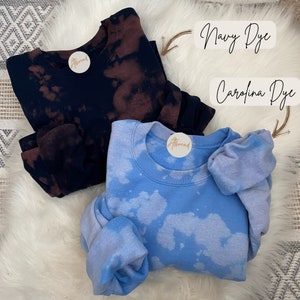May include: Two crewneck sweatshirts, one navy blue with brown bleach spots and one light blue with white bleach spots. The text "Navy Dye" and "Carolina Dye" are written above the sweatshirts. Both sweatshirts have a round sticker on the collar.