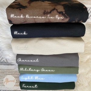 May include: A selection of folded fabric in shades such as black reverse tie dye, black, sand, charcoal, military green, light blue, and forest green. Each piece is labelled with handwritten text.