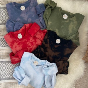May include: Five crewneck sweatshirts in shades of blue, red, black, green, and light blue, each with a tie-dye effect. The sweatshirts have a round label and are arranged on a fluffy white surface. The tie-dye effect gives each sweatshirt a unique look.
