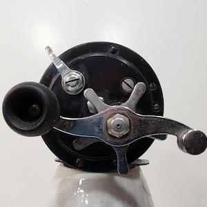 May include: A black fishing reel featuring a chrome-plated star drag handle and a black cylindrical knob. The circular side plate is secured with visible screws and includes a small chrome lever positioned at the top left of the central mechanism.